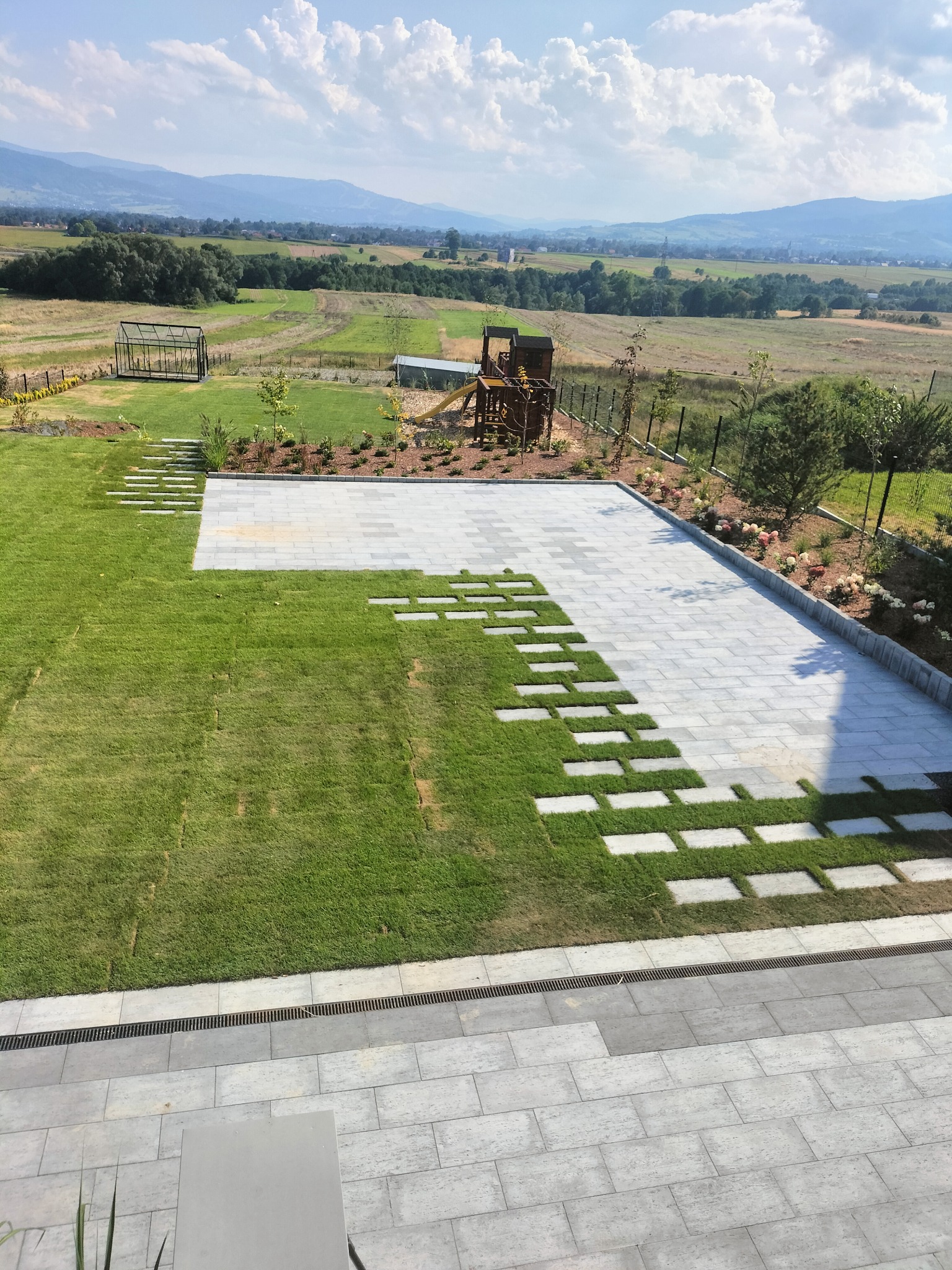 Widok z góry na ułożoną kostkę brukową na podjeździe, z elementami trawnika wkomponowanymi w wzór, w tle zielony krajobraz z domkiem ogrodowym i placem zabaw.