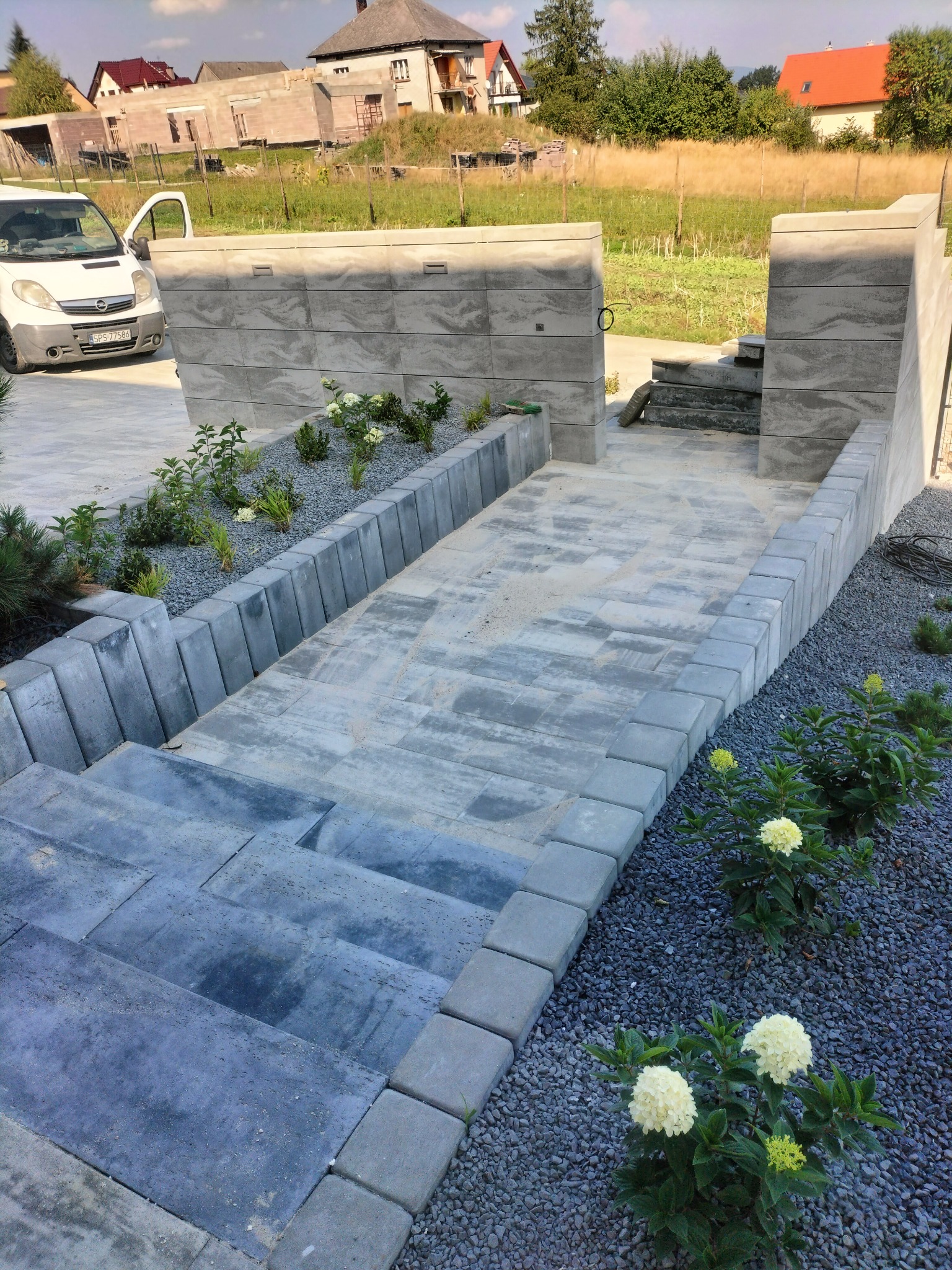 Ułożona ścieżka z kostki brukowej prowadząca do furtki, obrzeża z kostki, po bokach rabaty z kruszywem i roślinami, w tle budowa domu i zaparkowany bus.