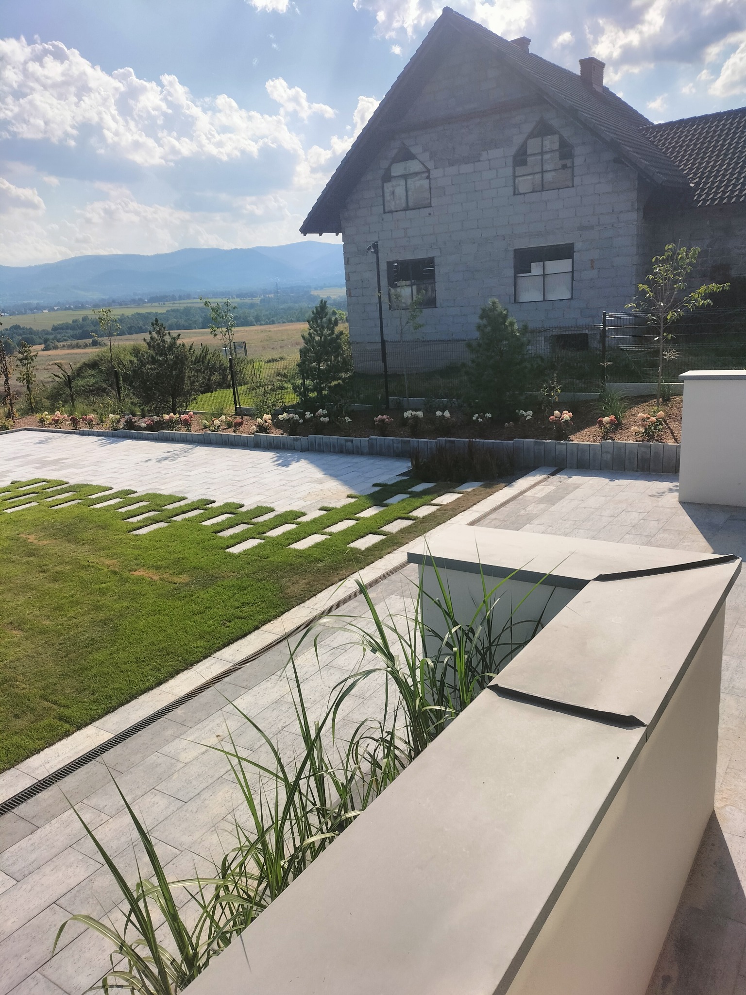 Nowoczesny podjazd z kostki brukowej przeplatany trawą, na tle domu w stanie surowym otoczonego zielenią i widokiem na góry.