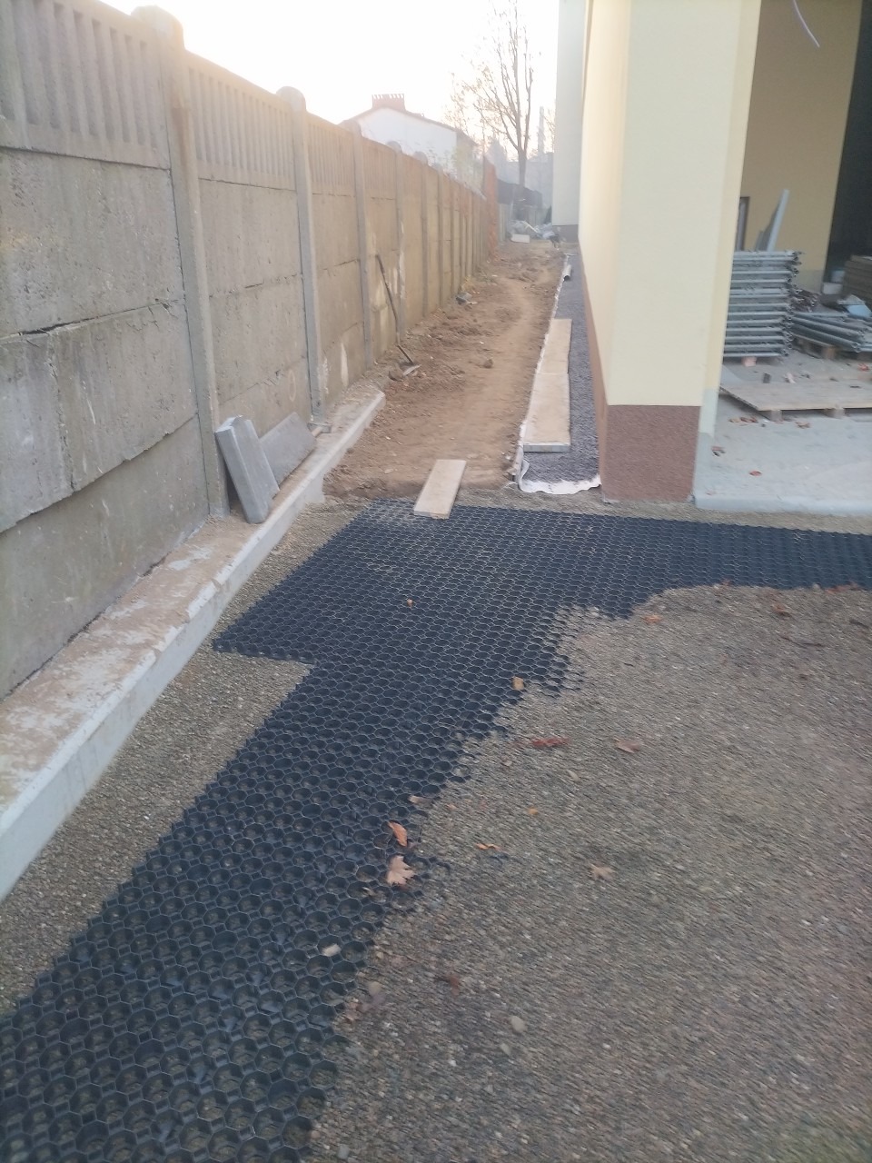 Przygotowanie podłoża pod nawierzchnię utwardzoną z wykorzystaniem geokraty, widoczny betonowy płot i fragment budynku w tle.