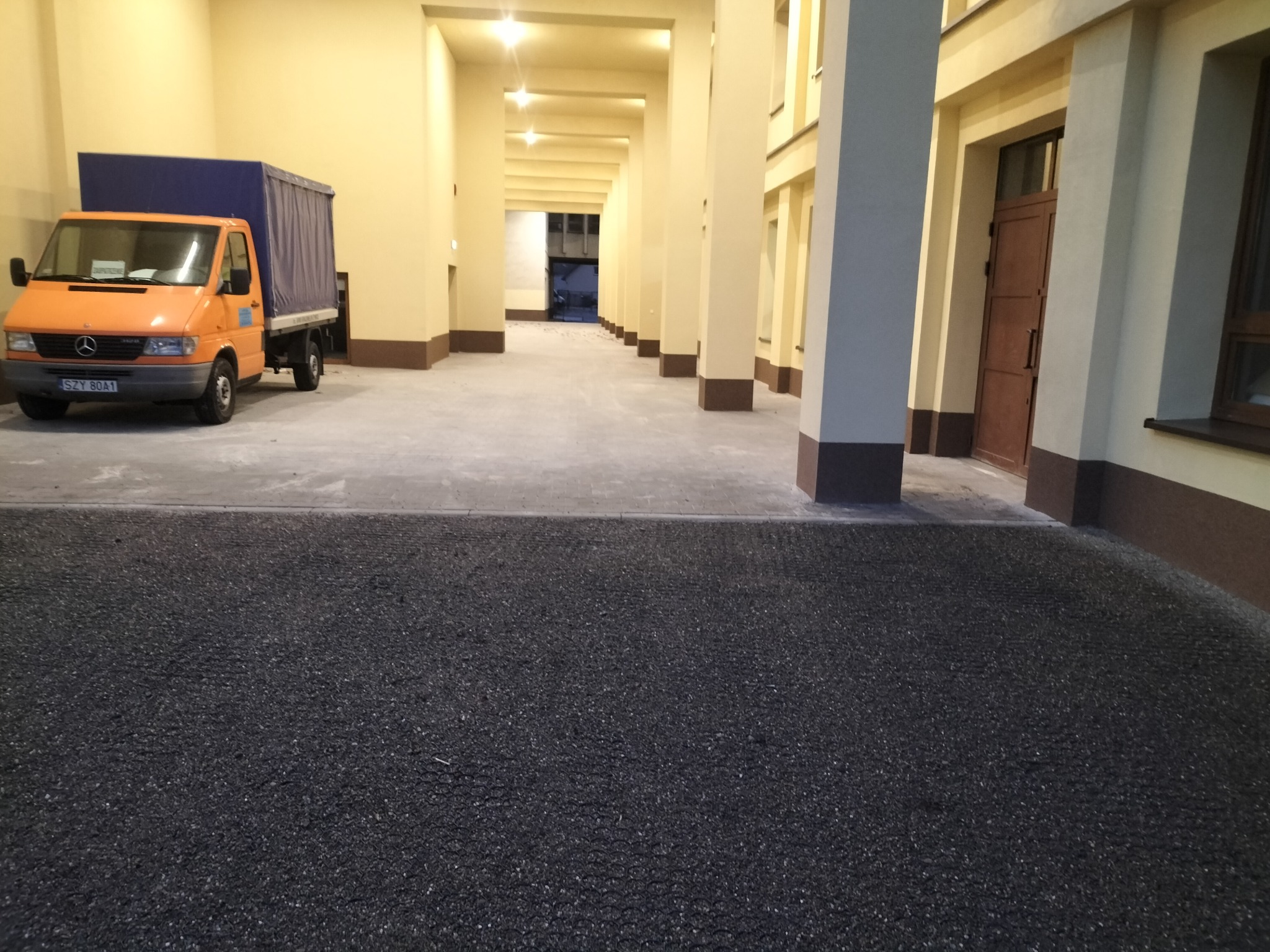 Pomarańczowy bus transportowy z niebieską plandeką zaparkowany na nowo ułożonej, ciemnej kostce brukowej na terenie hali z kolumnami.