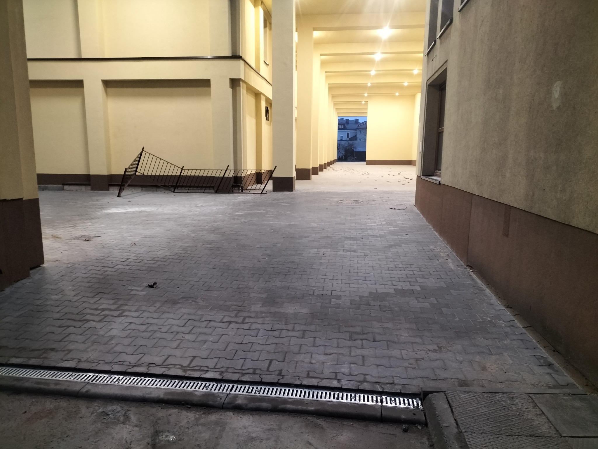 Szara kostka brukowa ułożona na dziedzińcu, z widoczną metalową kratką odpływową, fragmentem przewróconego metalowego ogrodzenia i kolumnadą w tle pod żółtym oświetleniem.