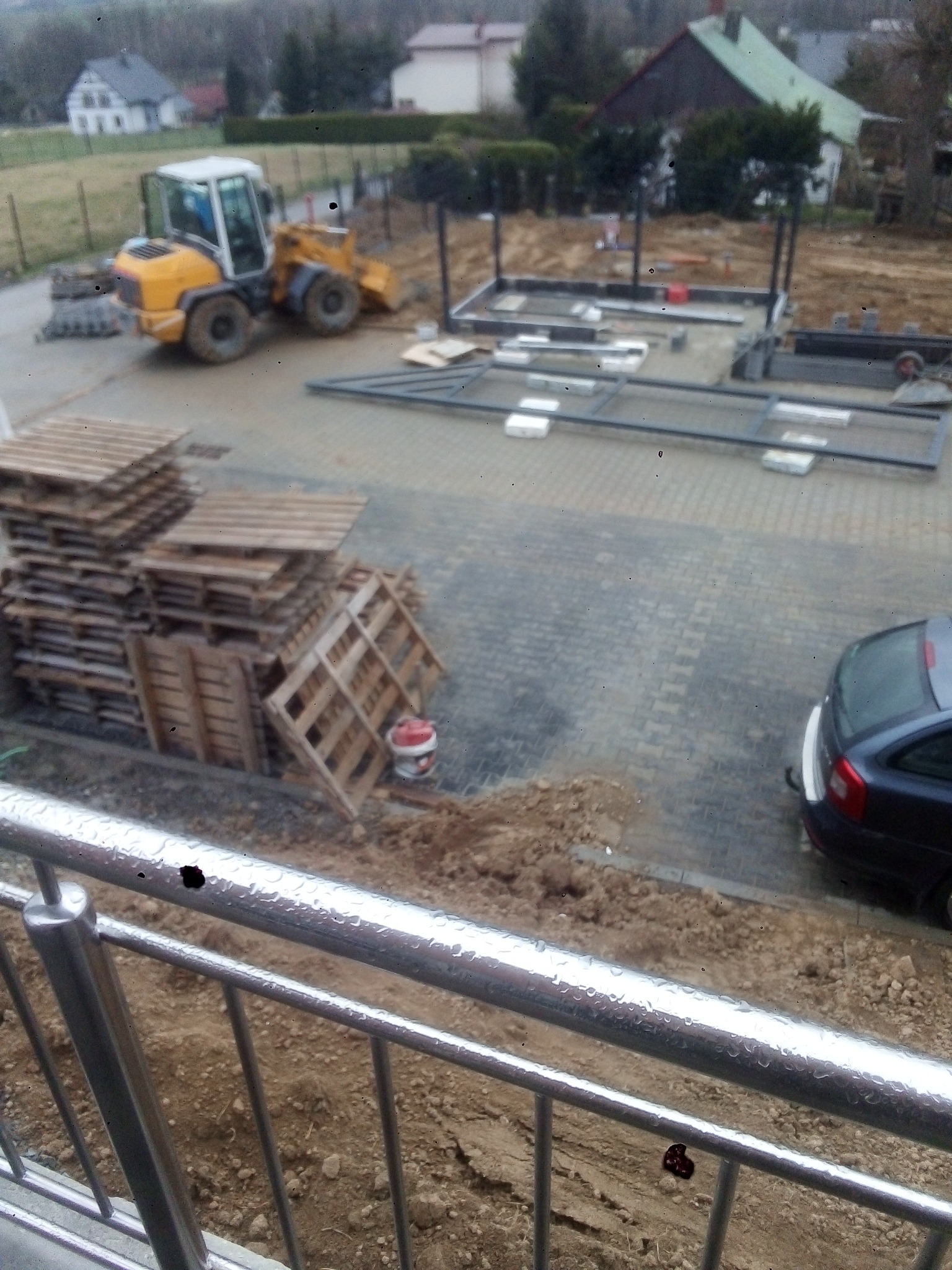 Widok z góry na plac budowy z paletami, traktorem i konstrukcją ogrodzenia w trakcie montażu na kostce brukowej, z perspektywy balkonu z metalową barierką.