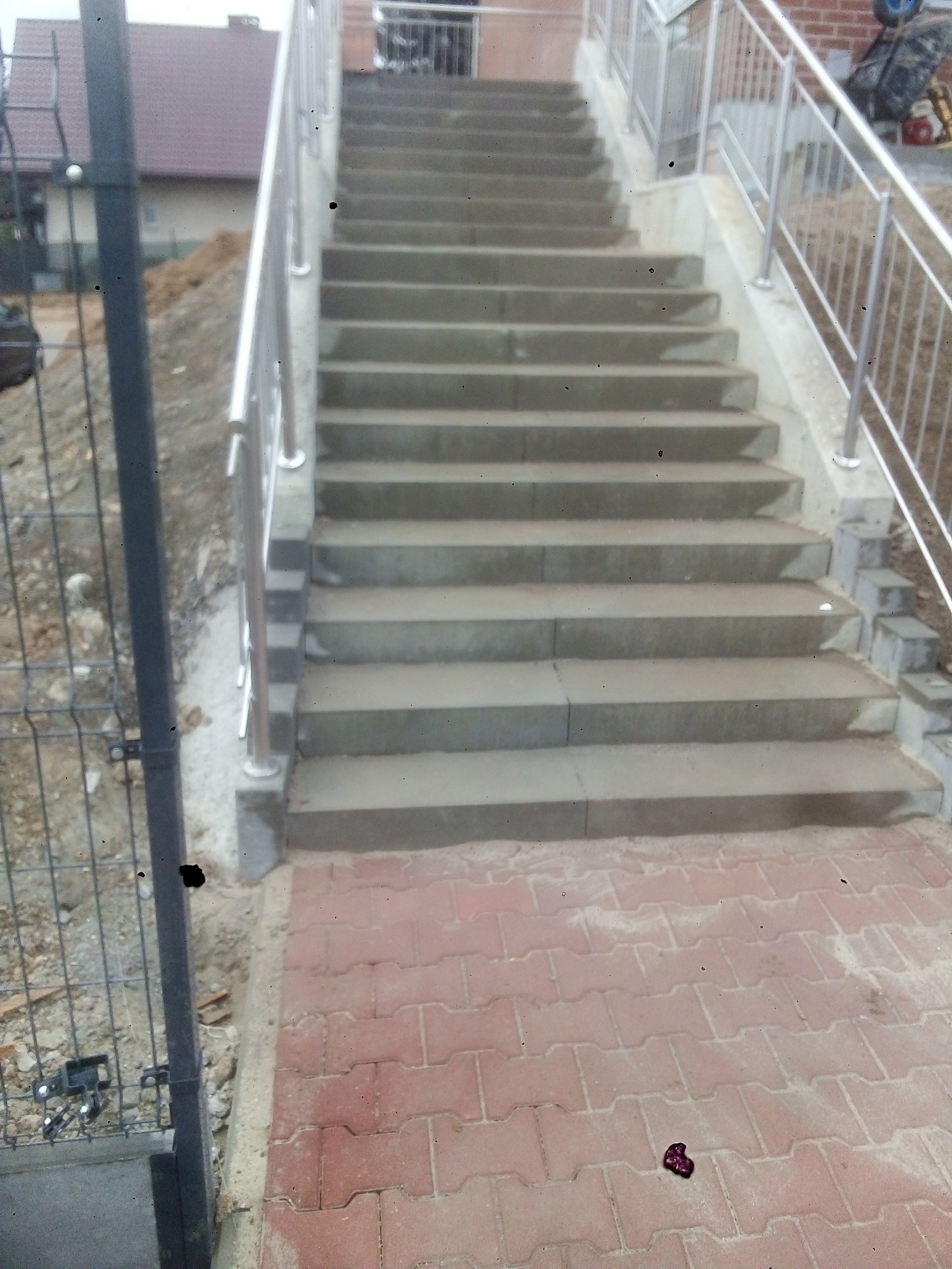 Betonowe schody zewnętrzne z metalową balustradą, prowadzące z chodnika z czerwonej kostki brukowej, obok ogrodzenia z siatki.