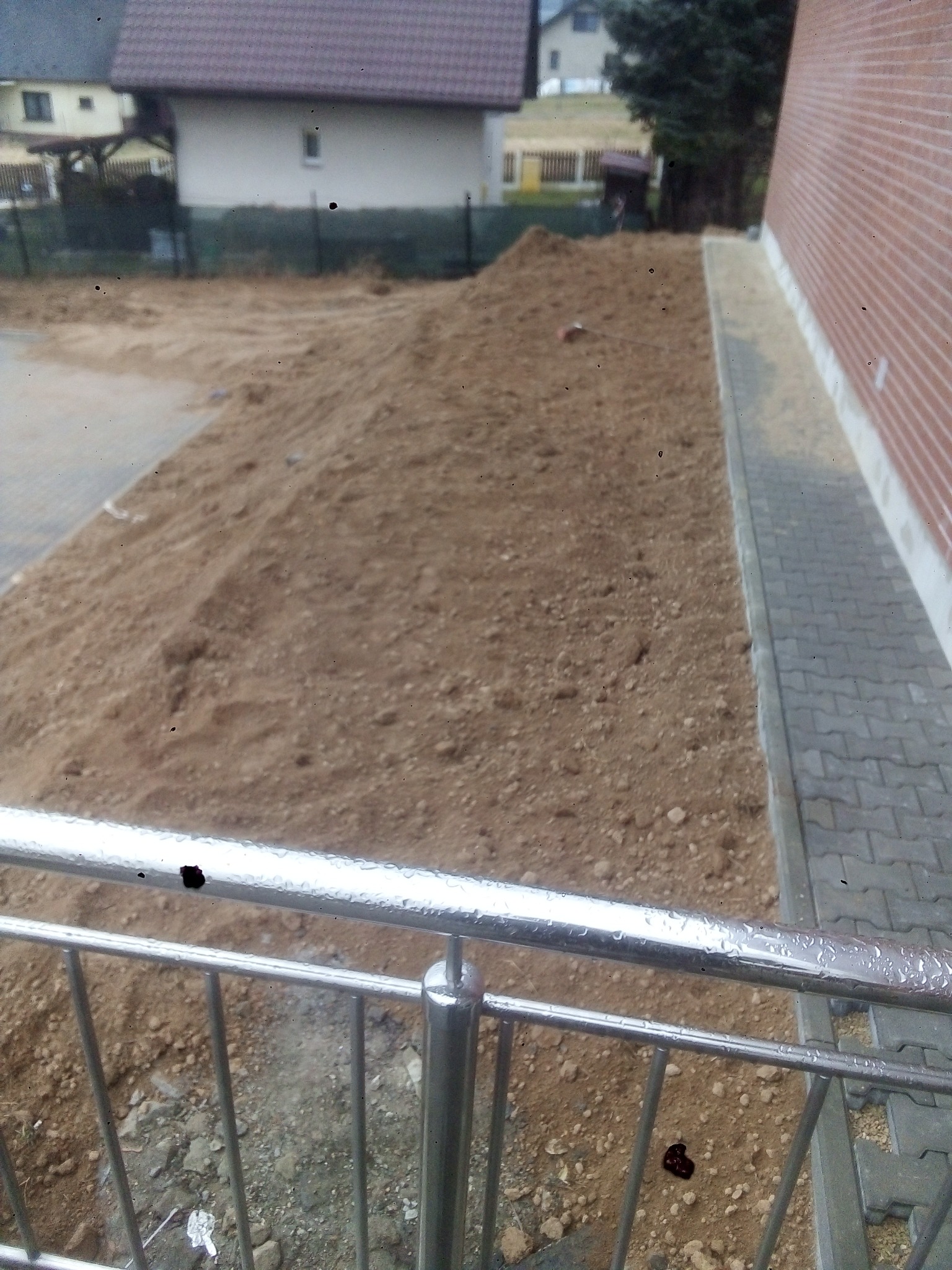 Nierówny teren z nasypem ziemi przy ścianie budynku z kostką brukową, widziany zza metalowego ogrodzenia.