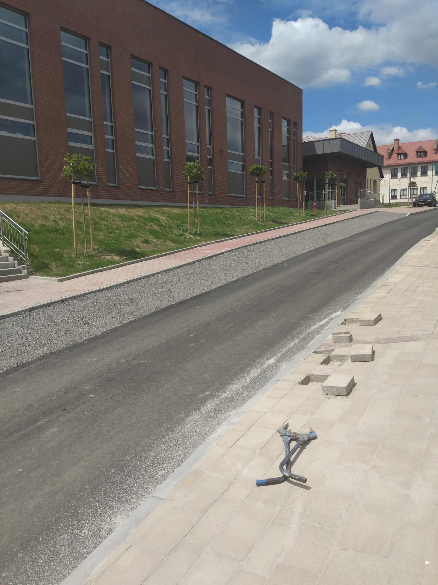 Nowa asfaltowa droga i chodnik z kostki brukowej w trakcie układania, z narzędziem brukarskim na pierwszym planie. Budynek w tle, Kalna.