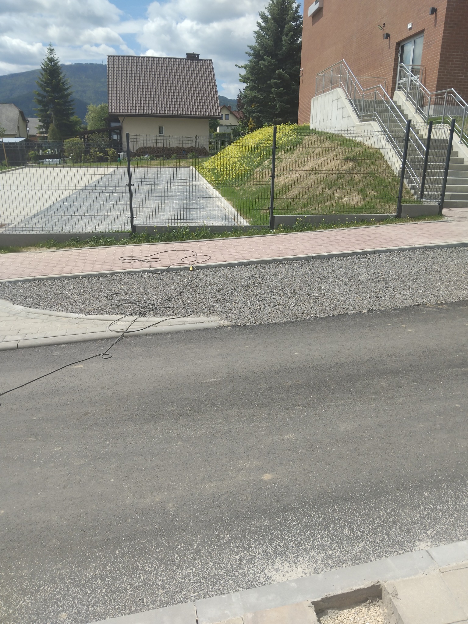 Wykończona asfaltowa droga z nowym chodnikiem z kostki, obok ogrodzenie panelowe. Widoczny kabel na żwirowym poboczu. W tle budynek i dom.