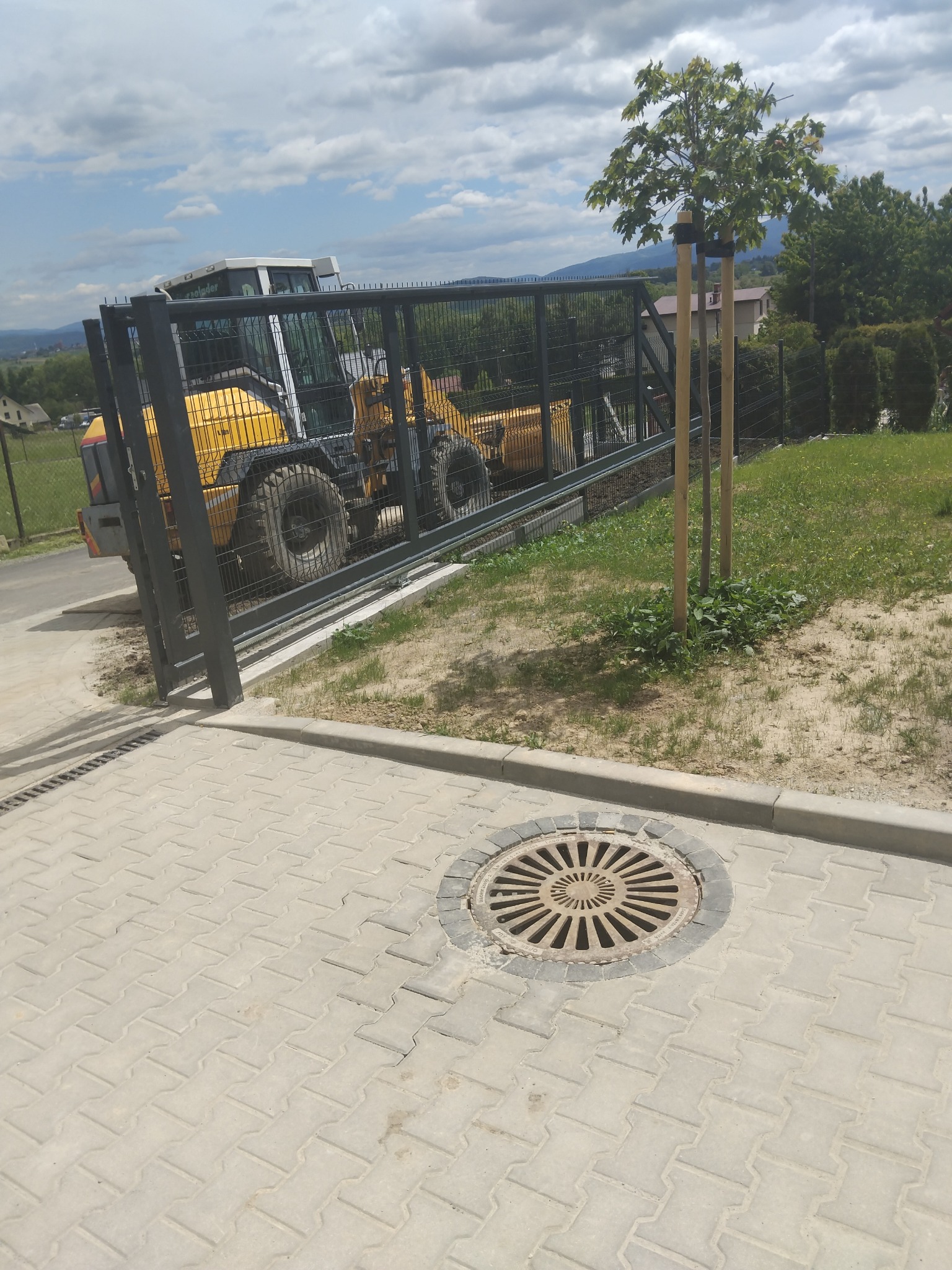 Żółta ładowarka za ogrodzeniem panelowym na kostce brukowej, z widocznym włazem kanalizacyjnym. W tle zieleń i pagórkowaty krajobraz, Kalna.