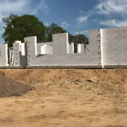 Wznoszone ściany parteru domu z białych bloczków na fundamencie, widoczny stos ziemi i drzewa w tle pod błękitnym niebem.