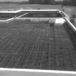 Zbrojenie płyty fundamentowej w szalunku z białego styropianu, widok z góry na siatkę z prętów stalowych, w tle wykopane fundamenty i krajobraz polny.