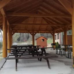 Drewniana altana z dachem i podłogą, wyposażona w stoły piknikowe, z widokiem na mały domek w tle.