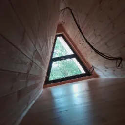 Wnętrze poddasza wykończonego drewnem, z trójkątnym oknem wychodzącym na zewnątrz i widocznymi kablami elektrycznymi.
