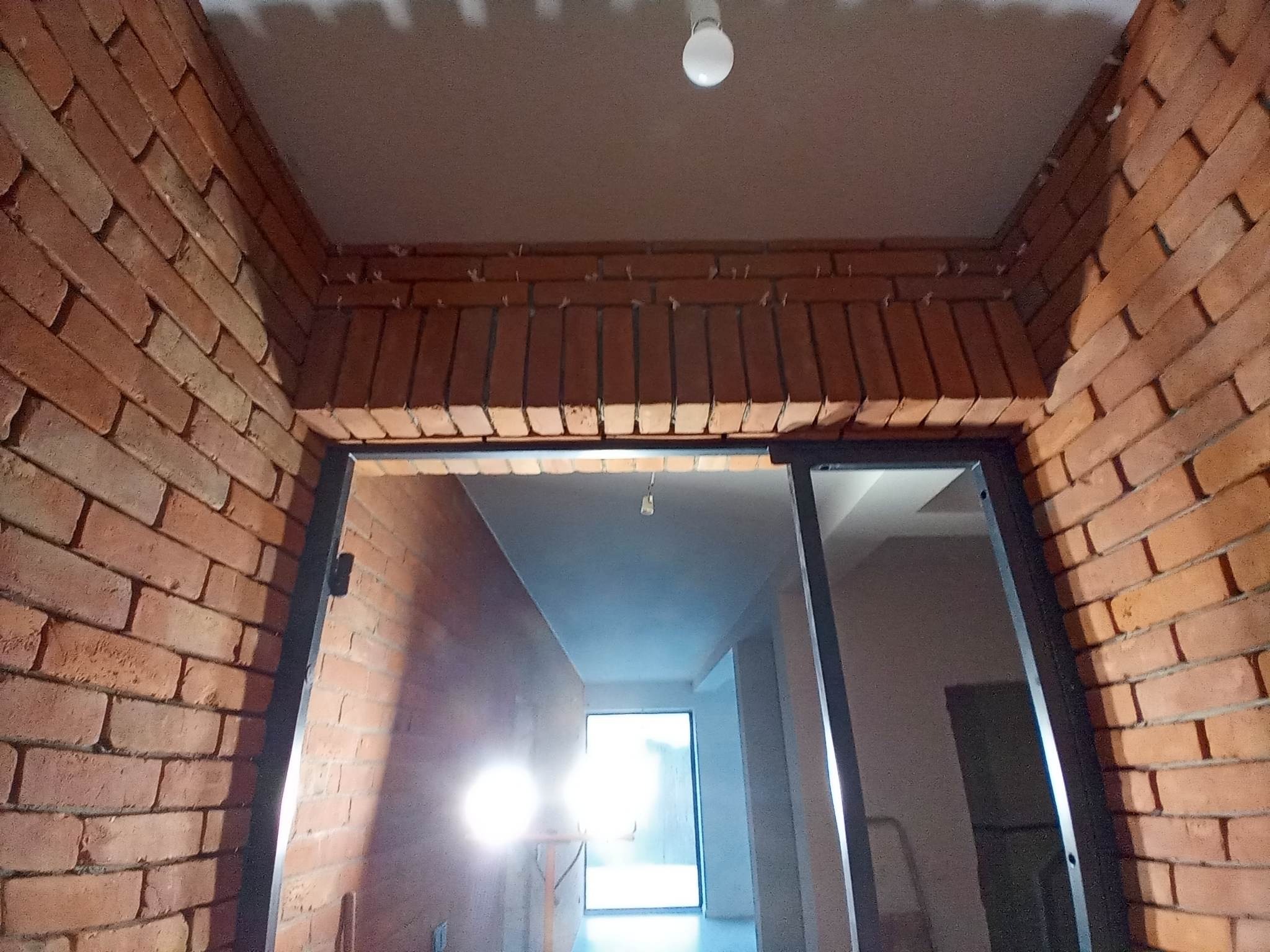 Surowe cegły tworzące łuk wejściowy z czarną ramą drzwiową wewnątrz budynku w trakcie budowy, widoczna żarówka na suficie i dalsza część korytarza z oknem.