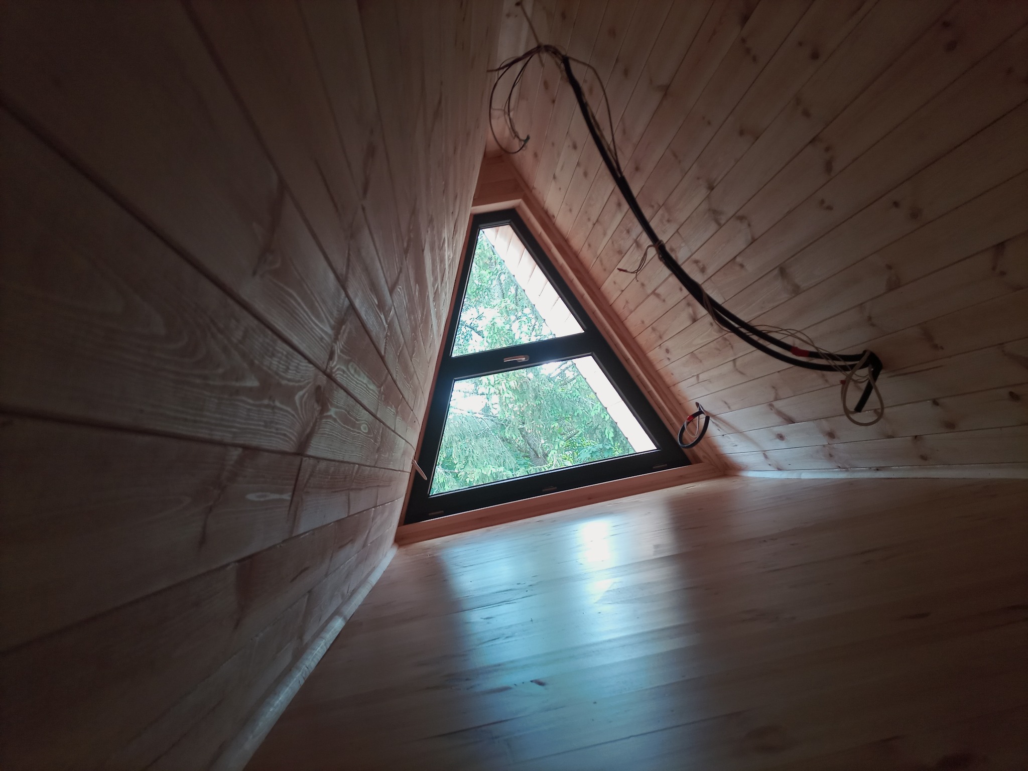 Wnętrze poddasza wykończonego drewnem, z trójkątnym oknem wychodzącym na zewnątrz i widocznymi kablami elektrycznymi.