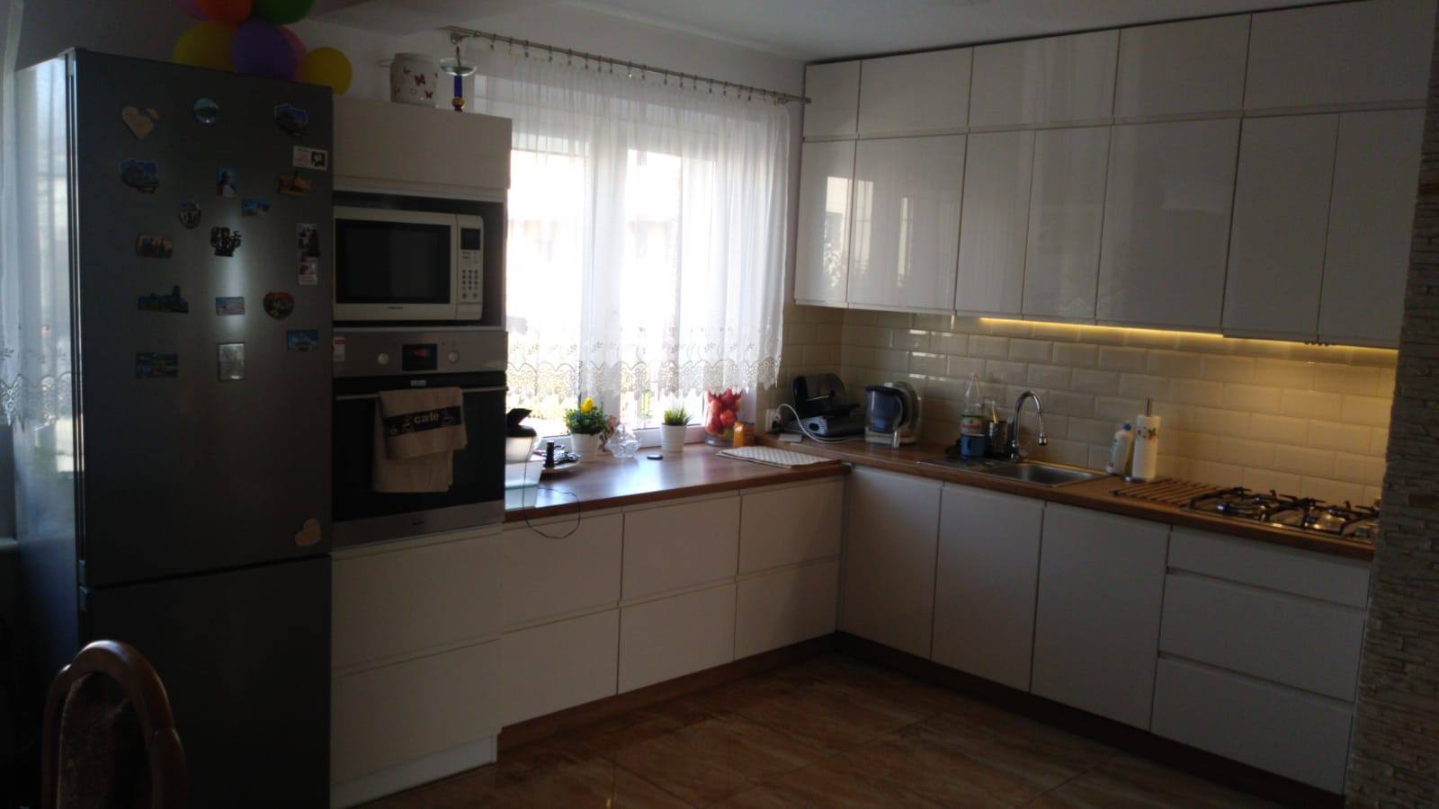 Jasna kuchnia z białymi, błyszczącymi frontami szafek, drewnianym blatem i oświetleniem LED pod szafkami wiszącymi. Na blacie kuchennym widoczne akcesoria kuchenne i dekoracje roślinne. Na lodówce...