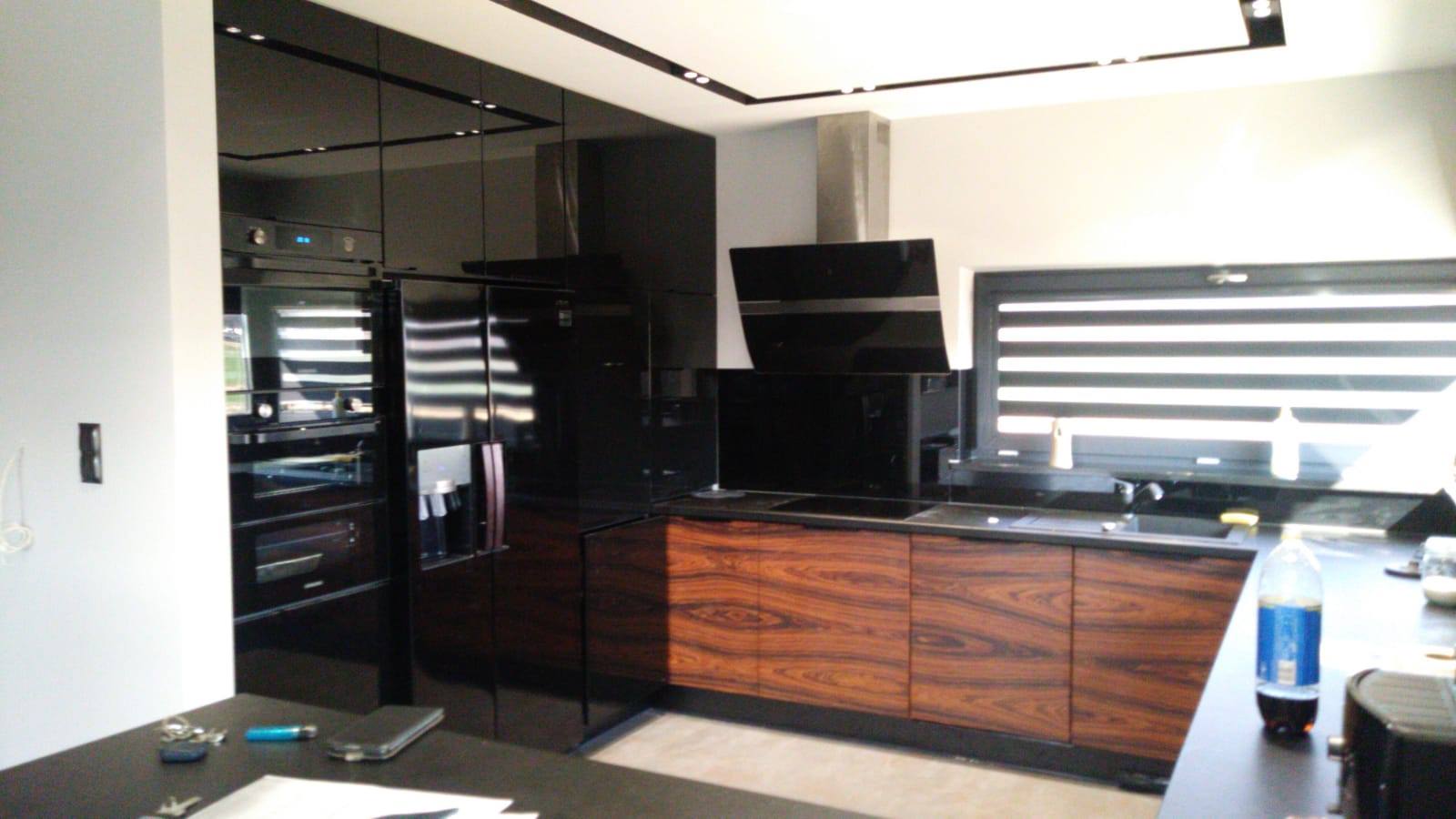 Nowoczesna kuchnia z czarnymi, lakierowanymi frontami szafek górnych i drewnianymi szafkami dolnymi z wyraźnym usłojeniem, widok na zabudowany piekarnik i lodówkę.