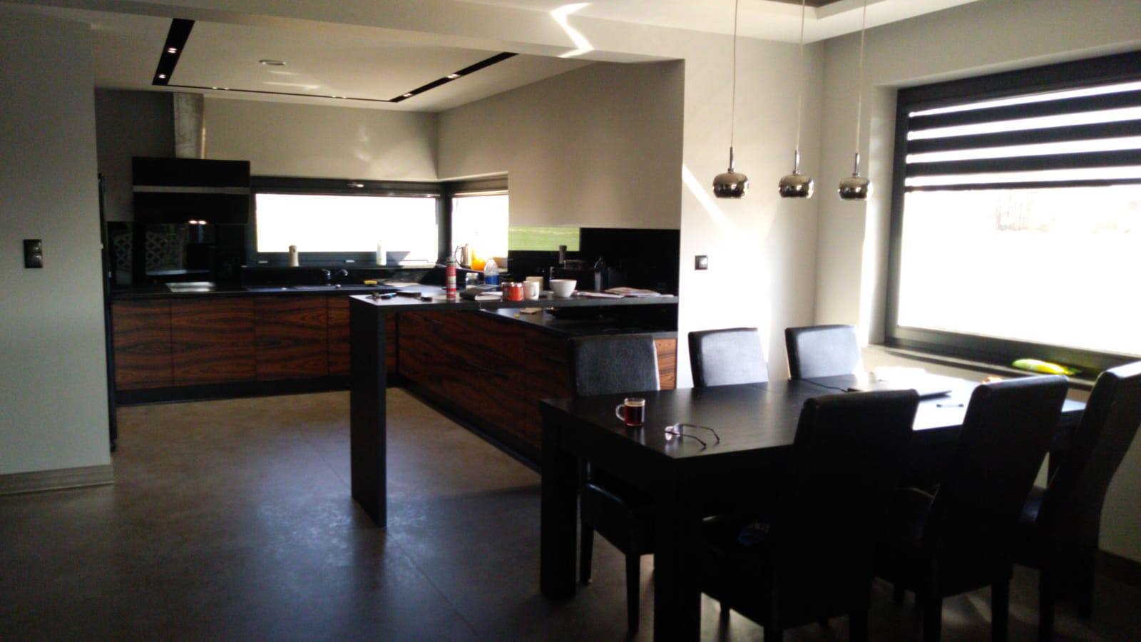 Nowoczesna kuchnia z wyspą i drewnianymi frontami szafek, połączona z jadalnią, widok z perspektywy stołu z filiżanką i okularami.