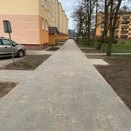 Długa, szara ścieżka z kostki brukowej prowadząca wzdłuż bloku mieszkalnego z beżowo-pomarańczową elewacją, z widocznym samochodem i drzewami w tle.