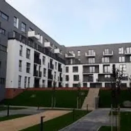 Nowoczesny budynek mieszkalny z białymi i szarymi elewacjami, balkonami i zagospodarowanym terenem zielonym z trawnikami i ścieżkami.