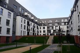 Nowoczesny budynek mieszkalny z białymi i szarymi elewacjami, balkonami i zagospodarowanym terenem zielonym z trawnikami i ścieżkami.