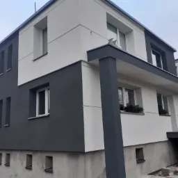 Dwukondygnacyjny dom z elewacją w kolorze białym i grafitowym, z widocznymi elementami konstrukcyjnymi i fragmentem odsłoniętego fundamentu, sugerującymi prace wykończeniowe.