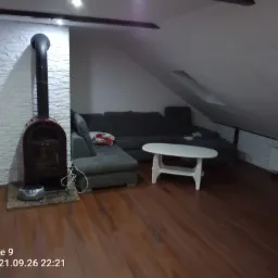 Salon na poddaszu z drewnianą podłogą, szarą sofą narożną, białym stolikiem kawowym i kominkiem obudowanym białą cegłą.