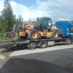 Pomarańczowa ładowarka teleskopowa na lawecie niebieskiej ciężarówki, gotowa do transportu. Widoczne drzewa i budynek w tle, słoneczny dzień.