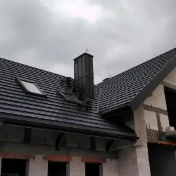 Nowy, czarny dach z dachówki modułowej, z oknem dachowym i kominem zwieńczonym wiatrowskazem w kształcie koguta, na surowym budynku z białego bloczka.