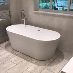 Nowoczesna wolnostojąca wanna owalna z chromowaną armaturą, umieszczona w łazience wykończonej płytkami w odcieniach szarości, z widokiem na zieleń przez okno.