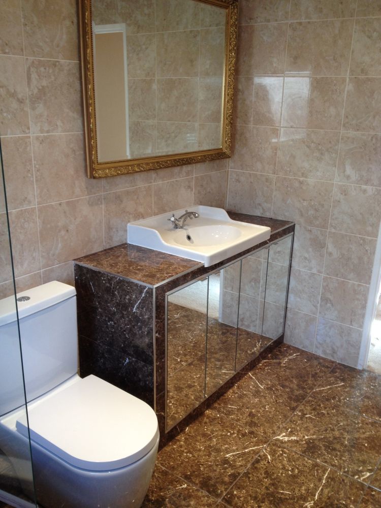 Wykończona łazienka z marmurową umywalką z szafką z lustrzanymi drzwiczkami, toaleta oraz duże, złote lustro na ścianie z beżowych płytek.