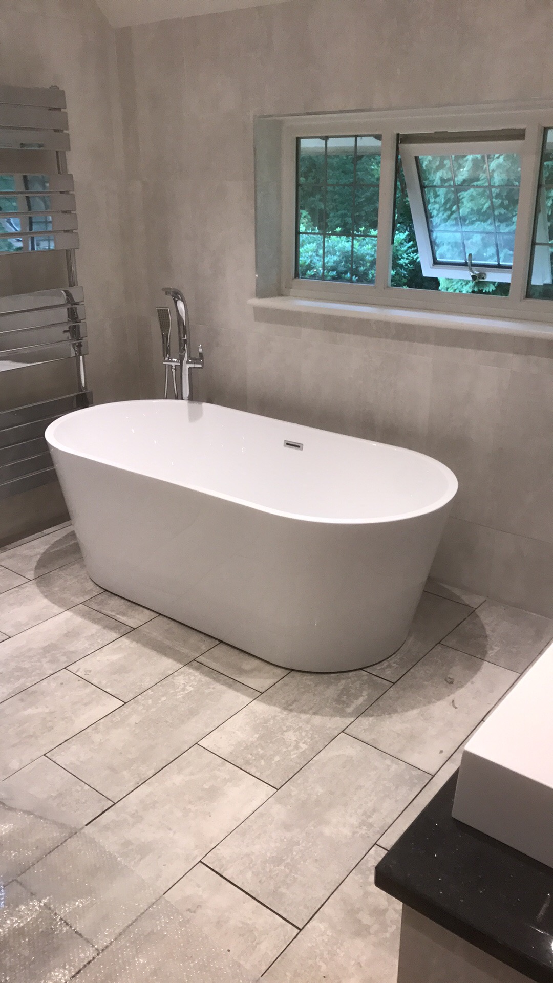 Nowoczesna wolnostojąca wanna owalna z chromowaną armaturą, umieszczona w łazience wykończonej płytkami w odcieniach szarości, z widokiem na zieleń przez okno.