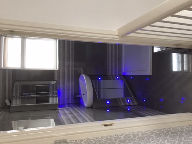 Nowoczesna łazienka z szarą tapetą w paski, podświetlanym na niebiesko lustrem i szafkami oraz oknem z białymi ramami.