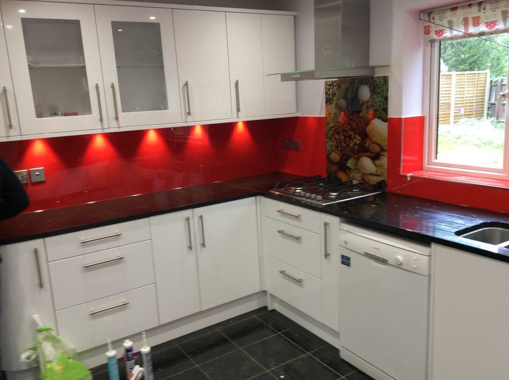Nowoczesna kuchnia z białymi szafkami, czarnym blatem i czerwonym panelem szklanym podświetlonym punktowo, widoczny fragment okna z roletą w filiżanki.