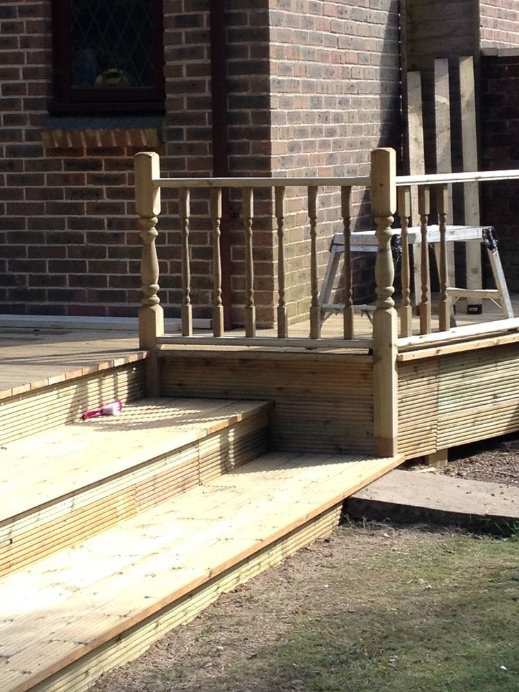 Nowo wybudowany drewniany taras z barierką i schodami, prowadzący do drzwi wejściowych domu z cegły, na trawniku leży różowa rolka taśmy.