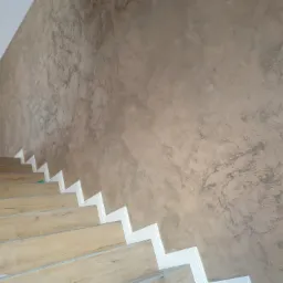 Ściana z dekoracyjną fakturą w odcieniach beżu i szarości, sąsiadująca z drewnianymi schodami z białymi listwami przypodłogowymi.