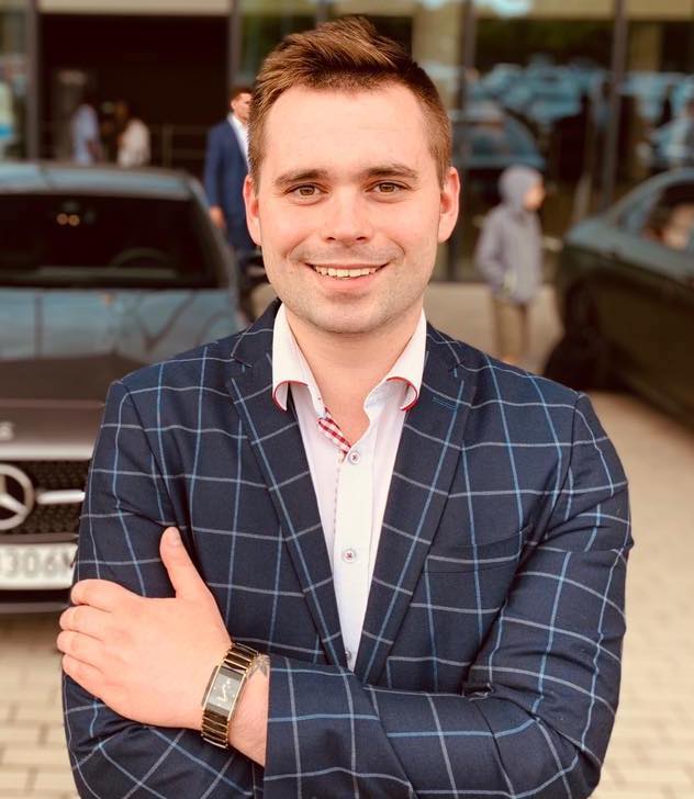 Uśmiechnięty mężczyzna w niebieskiej marynarce w kratę, białej koszuli i z zegarkiem na tle samochodów i budynku z przeszkloną fasadą.