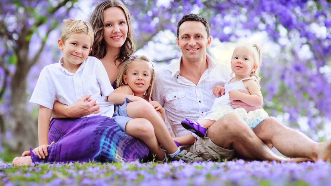 Uśmiechnięta rodzina z trójką dzieci siedzi na trawie usianej fioletowymi kwiatami, w tle kwitnące drzewo.