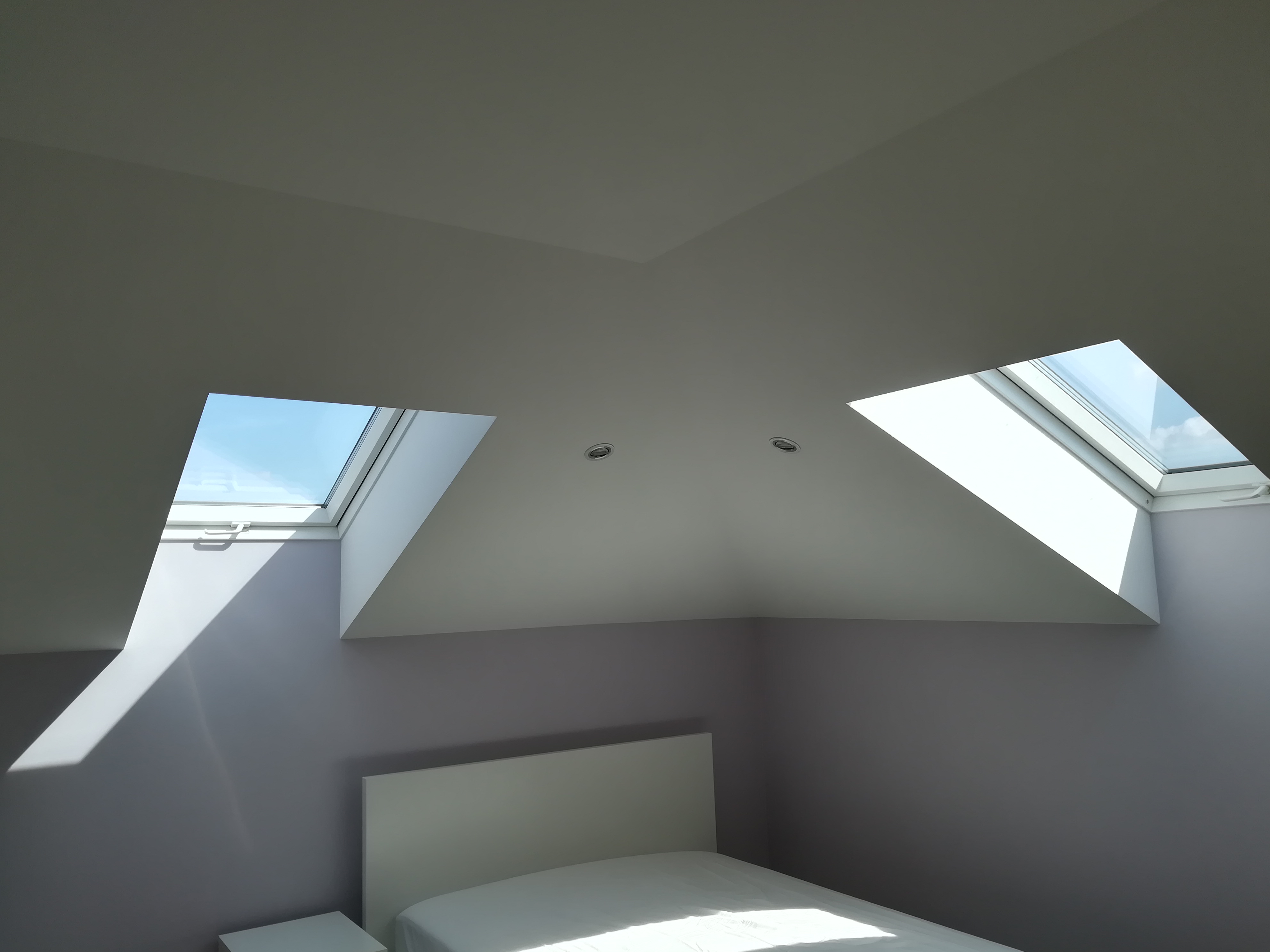 Pokój na poddaszu z dwoma oknami dachowymi, pomalowanymi ścianami na szaro, widoczne oświetlenie punktowe w suficie oraz fragment łóżka z białym zagłówkiem i pościelą.