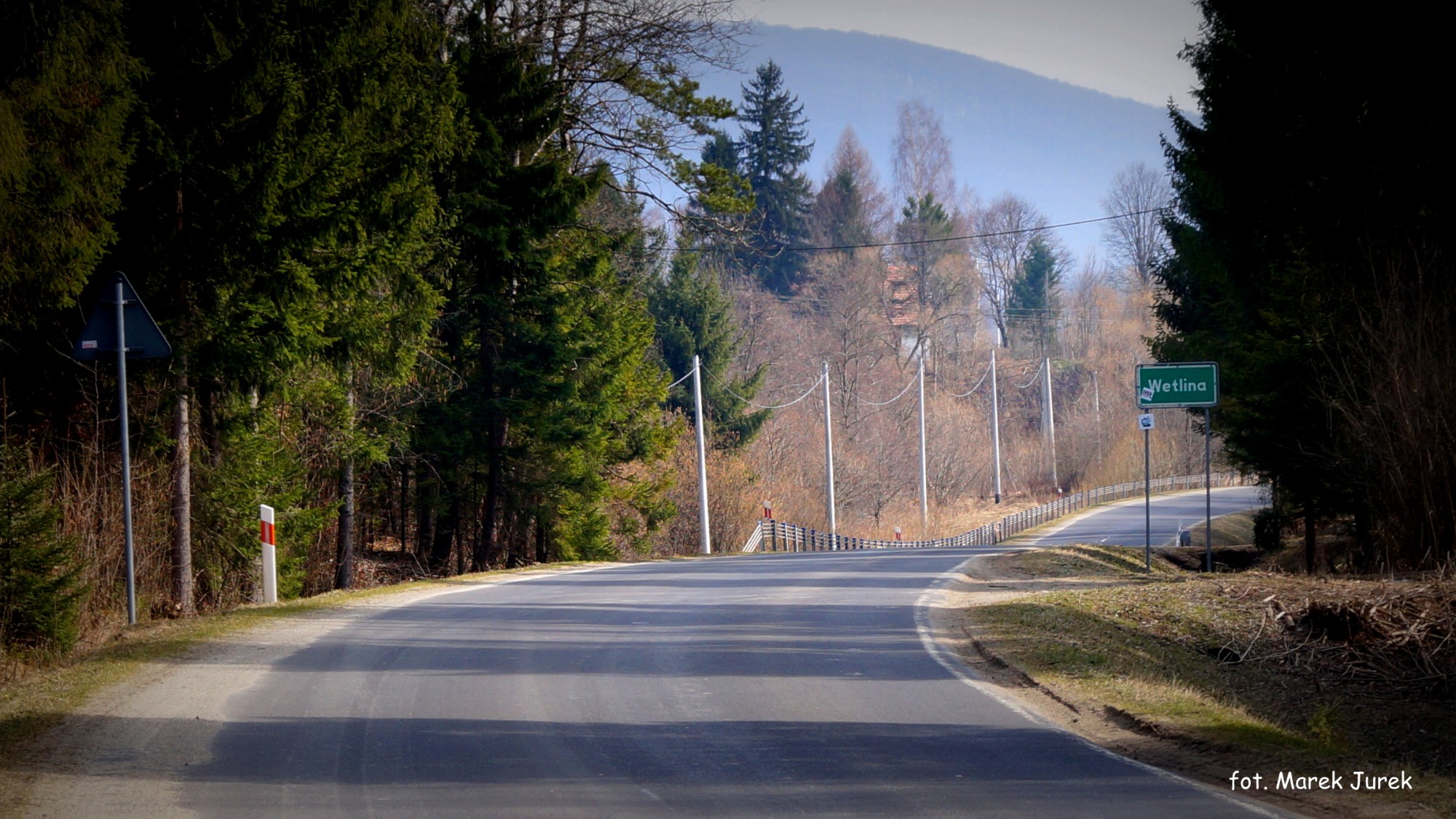 Asfaltowa droga wijąca się przez las w kierunku Wetliny, z widocznym znakiem miejscowości i górą w tle.