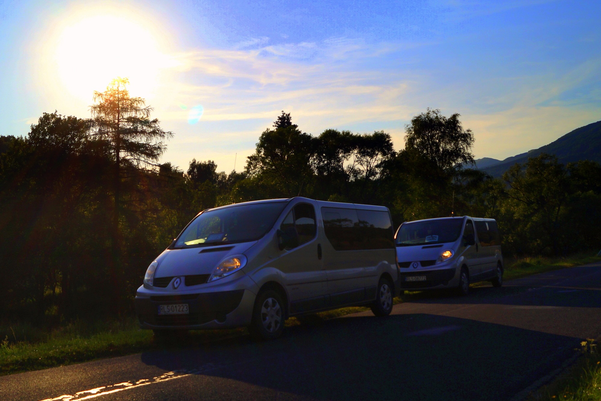 Dwa srebrne busy Renault Trafic zaparkowane na asfalcie na tle drzew i zachodzącego słońca, widoczne numery rejestracyjne PLS 01223 i PLS 89772.