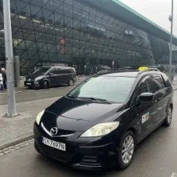 Czarny samochód typu minivan, oznaczony jako taksówka z numerem bocznym 9373, zaparkowany przed nowoczesnym budynkiem lotniska z przeszkloną fasadą o strukturze kratownicy.