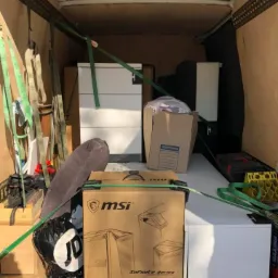 Wnętrze furgonetki załadowane kartonami, meblami i workami zabezpieczonymi zielonymi pasami transportowymi, widoczny karton z grafiką komputera stacjonarnego MSI Infinite Series.