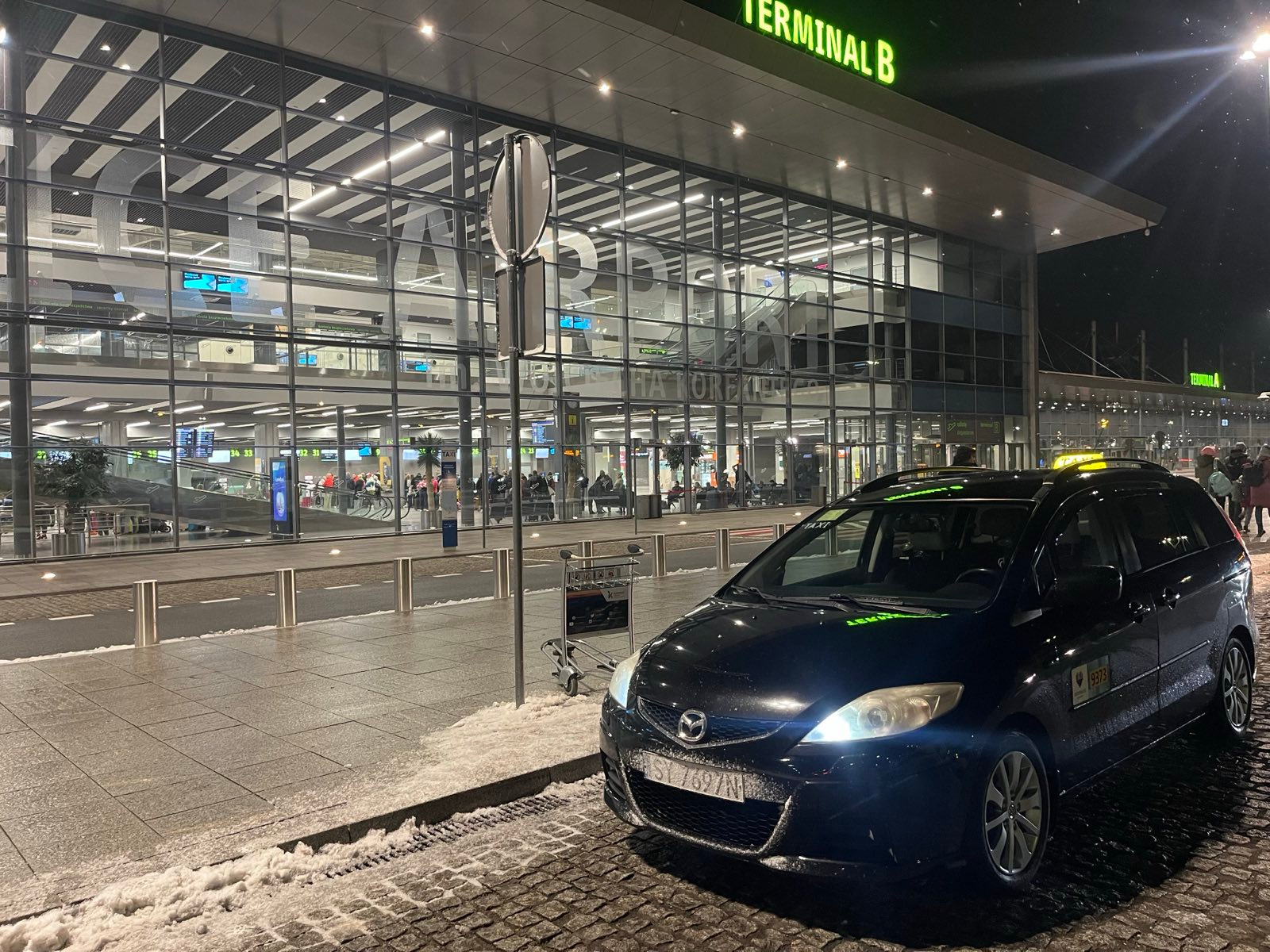 Czarny samochód typu van z żółtym znakiem taxi na dachu, zaparkowany przed oświetlonym budynkiem terminalu lotniska w zimowy wieczór.