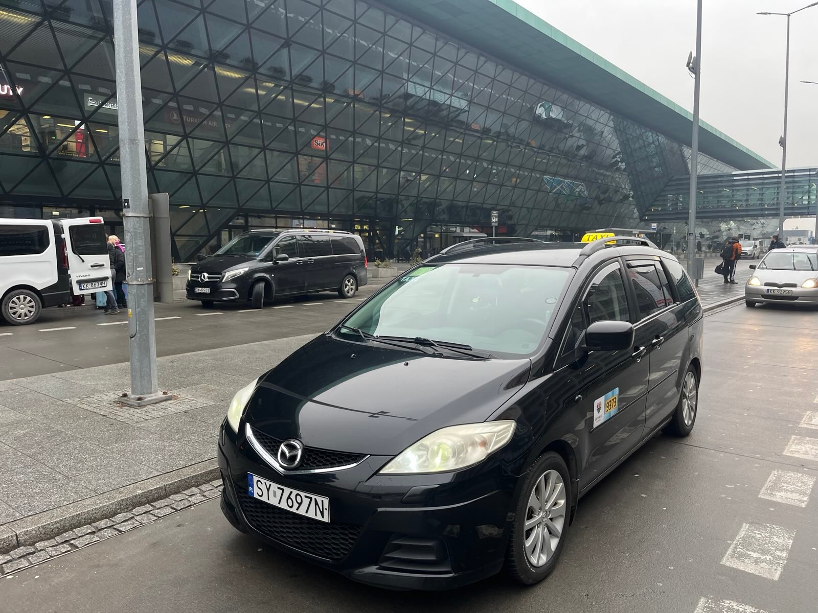 Czarny samochód typu minivan, oznaczony jako taksówka z numerem bocznym 9373, zaparkowany przed nowoczesnym budynkiem lotniska z przeszkloną fasadą o strukturze kratownicy.