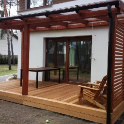 Nowoczesny taras z drewna z pergolą, przytulny kącik z drewnianymi krzesłami i stołem, widok na dom i ogród, zieleń trawnika i drzew w tle.