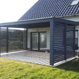 Nowoczesna, szara pergola z poziomymi listwami, przylegająca do białego domu z ciemnym dachem, rzucająca cienie na jasny taras i trawnik.