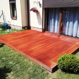 Czerwono-brązowy taras z desek, przylegający do domu z białymi ścianami i zwiniętą roletą okienną, otoczony trawnikiem i krzewami.