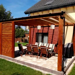 Drewniana konstrukcja z zadaszeniem i lamelami, tworząca zacienione miejsce do siedzenia z widokiem na trawnik; stół z krzesłami, oświetlenie wiszące, obok domu z ceglaną ścianą.