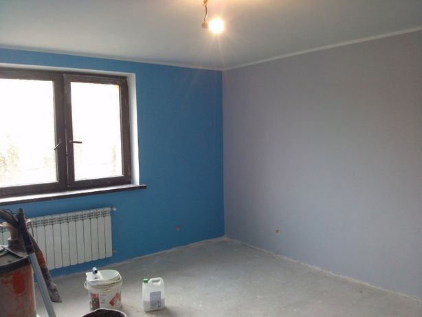 Pokój po malowaniu ścian na dwa kolory: niebieski przy oknie i szary na pozostałych ścianach, widoczne wiaderka z farbą i grzejnik pod oknem.