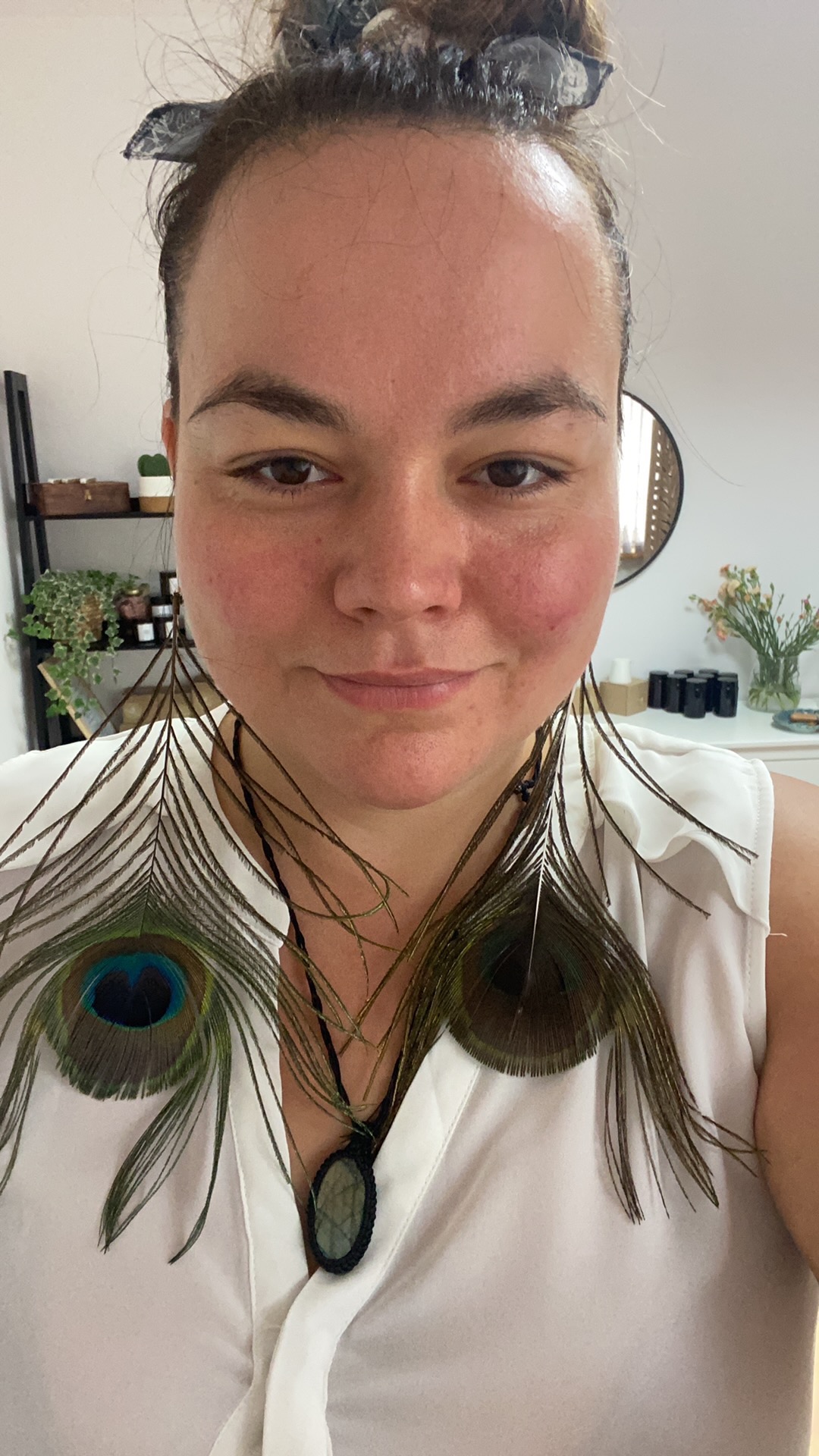 Portret kobiety z kolczykami z pawich piór i naszyjnikiem z kamieniem, uśmiechająca się do kamery w jasnym wnętrzu.